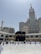 A large gathering of people surrounding the Kaaba in Mecca, with the Abraj Al-Bait Clock Tower and other skyscrapers in the background. The courtyard is spacious and bright, filled with individuals dressed in traditional white garments, denoting a religious pilgrimage.