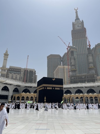 A large gathering of people surrounding the Kaaba in Mecca, with the Abraj Al-Bait Clock Tower and other skyscrapers in the background. The courtyard is spacious and bright, filled with individuals dressed in traditional white garments, denoting a religious pilgrimage.