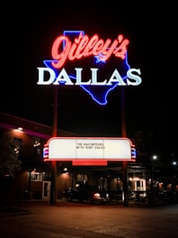 A bright neon sign featuring the name 'Gilley's' in red cursive above 'DALLAS' in white block letters, outlined in blue in the shape of Texas. A marquee below displays 'The Raconteurs with Hunt Sales' and the website 'www.gilleysdallas.com'. The scene is set at night with warm outdoor lighting illuminating the brick building and a parked vehicle nearby.