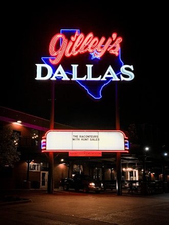 A bright neon sign featuring the name 'Gilley's' in red cursive above 'DALLAS' in white block letters, outlined in blue in the shape of Texas. A marquee below displays 'The Raconteurs with Hunt Sales' and the website 'www.gilleysdallas.com'. The scene is set at night with warm outdoor lighting illuminating the brick building and a parked vehicle nearby.