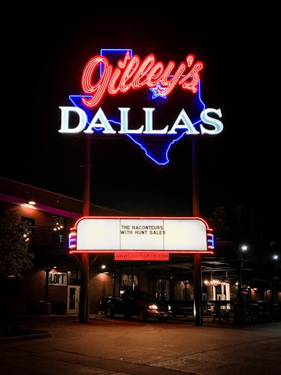 A bright neon sign featuring the name 'Gilley's' in red cursive above 'DALLAS' in white block letters, outlined in blue in the shape of Texas. A marquee below displays 'The Raconteurs with Hunt Sales' and the website 'www.gilleysdallas.com'. The scene is set at night with warm outdoor lighting illuminating the brick building and a parked vehicle nearby.