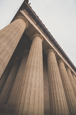 A dramatic perspective of ancient-style stone columns with fluted shafts supporting a large architectural structure. The columns ascend towards an ornately detailed roofline, evoking a sense of grandeur and historical significance. The muted lighting adds a timeless atmosphere.