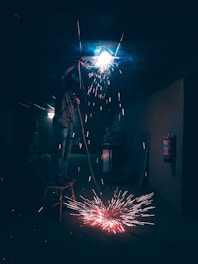A person wearing protective gear stands on a chair in a dimly lit room, performing welding work with bright sparks flying from the welding point. The room has a dark, industrial feel with visible scattered equipment and a fire extinguisher mounted on the wall.