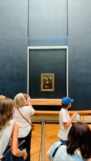 A group of people are gathered in front of a famous painting that is displayed behind glass in a museum. The painting is enclosed in a large, ornate frame, and it attracts significant attention from visitors, some of whom are taking photos.