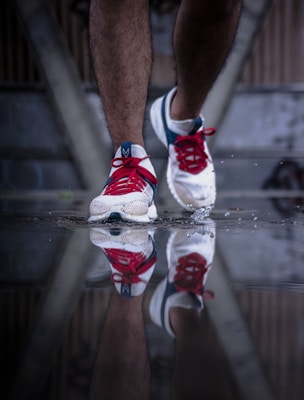 A close-up of a person wearing white running shoes with red and blue accents. The shoes are partially submerged in water, creating a clear reflection on the wet surface.