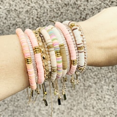 A forearm adorned with multiple beaded and metallic bracelets in various shades, including pink, white, and gold. The accessories feature different textures and designs, such as round beads and metallic elements, arranged harmoniously across the wrist.