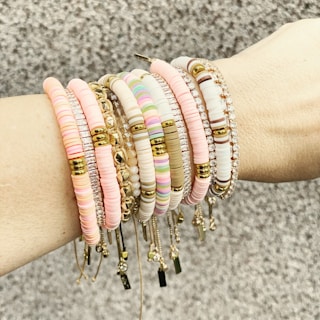 A forearm adorned with multiple beaded and metallic bracelets in various shades, including pink, white, and gold. The accessories feature different textures and designs, such as round beads and metallic elements, arranged harmoniously across the wrist.
