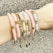 A forearm adorned with multiple beaded and metallic bracelets in various shades, including pink, white, and gold. The accessories feature different textures and designs, such as round beads and metallic elements, arranged harmoniously across the wrist.