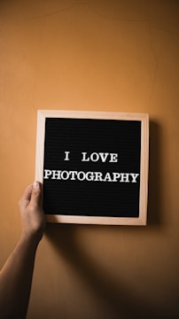 A hand is holding a wooden-framed letter board with the text 'I LOVE PHOTOGRAPHY' in white capital letters against a black background. The background is a plain, light brown wall.