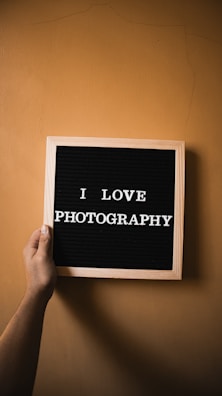A hand is holding a wooden-framed letter board with the text 'I LOVE PHOTOGRAPHY' in white capital letters against a black background. The background is a plain, light brown wall.