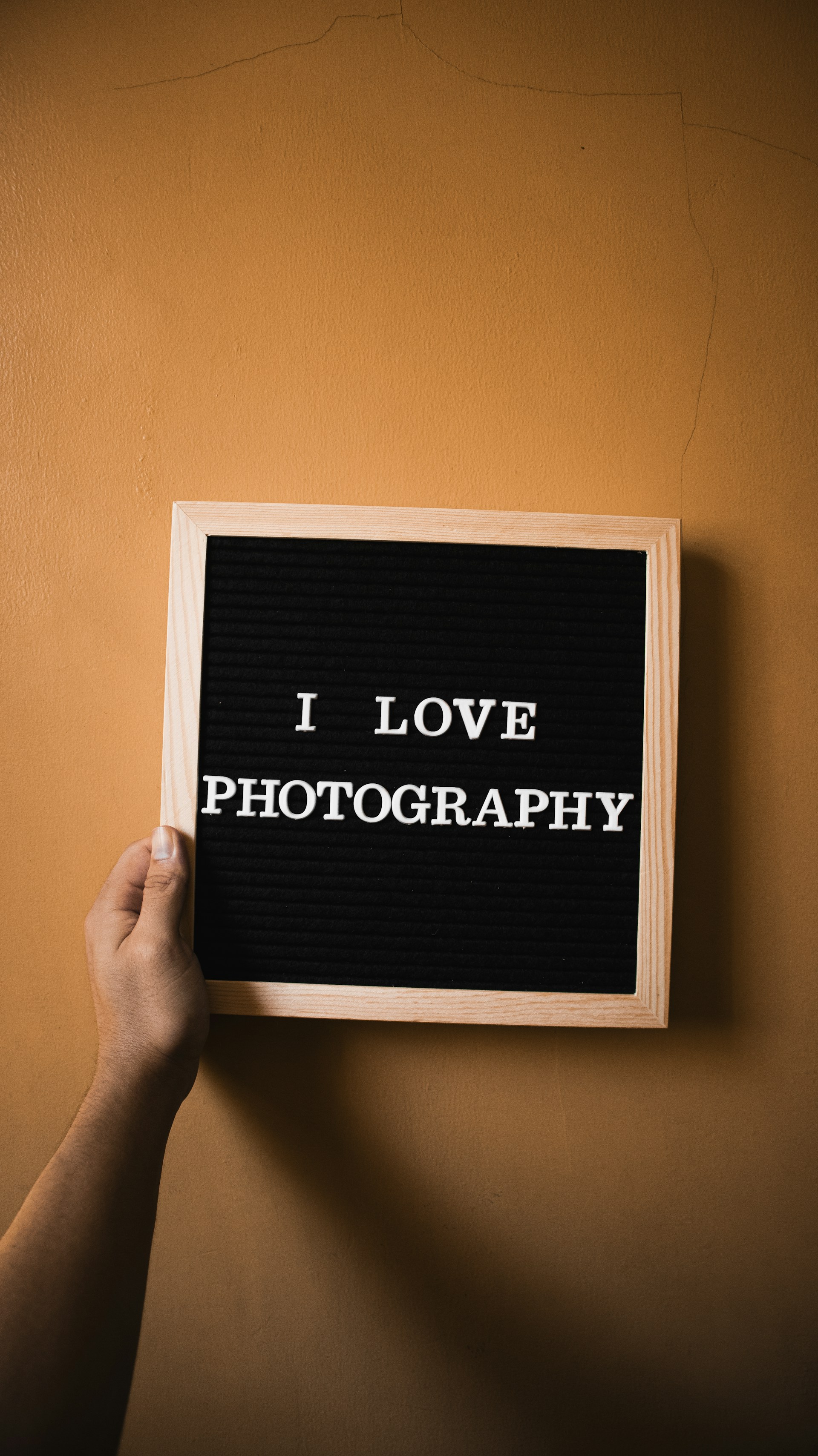 A hand is holding a wooden-framed letter board with the text 'I LOVE PHOTOGRAPHY' in white capital letters against a black background. The background is a plain, light brown wall.