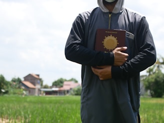 A person is standing outdoors in a field holding a book with a prominent gold emblem on the cover. The individual is wearing a dark hooded garment and a face mask, suggesting a certain level of anonymity. In the background, there are some blurred residential buildings, along with green grass and trees, under a partly cloudy sky.