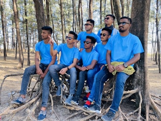A group of seven young men wearing matching blue shirts and jeans are sitting and standing around large tree roots in a forest area. They are all wearing sunglasses and appear to be relaxed, some looking into the distance.