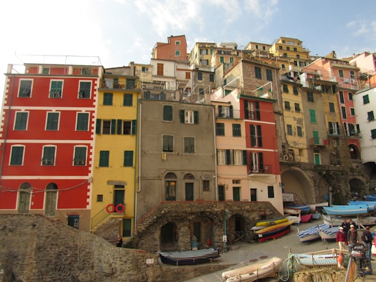 A vibrant coastal village with multicolored buildings stacked closely together, showcasing hues of red, yellow, orange, and peach. The architecture is traditional with verdant shutters and a rustic vibe. Small boats are docked along the waterfront pathway, and a few people are walking, enjoying the picturesque surroundings.