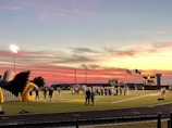 A sports field with a football game unfolding under a vibrant sunset sky, featuring a large inflatable mascot and a scoreboard displaying the score. Players and coaches are visible on the green field, surrounded by a track and some flags waving in the breeze.