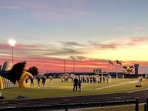 A sports field with a football game unfolding under a vibrant sunset sky, featuring a large inflatable mascot and a scoreboard displaying the score. Players and coaches are visible on the green field, surrounded by a track and some flags waving in the breeze.