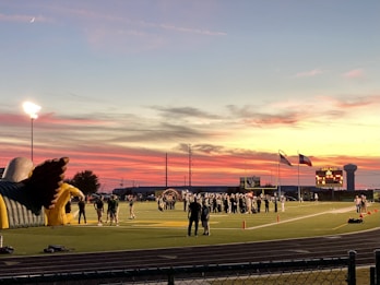 A sports field with a football game unfolding under a vibrant sunset sky, featuring a large inflatable mascot and a scoreboard displaying the score. Players and coaches are visible on the green field, surrounded by a track and some flags waving in the breeze.