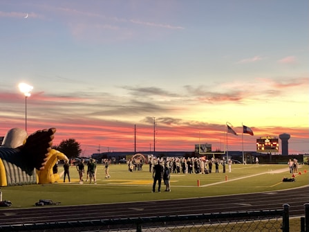 A sports field with a football game unfolding under a vibrant sunset sky, featuring a large inflatable mascot and a scoreboard displaying the score. Players and coaches are visible on the green field, surrounded by a track and some flags waving in the breeze.