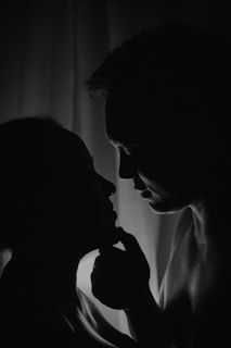 A black and white silhouette of two people facing each other closely. One person gently holds the other's chin, creating an intimate and tender moment. The background features soft, diffused lighting, enhancing the scene's depth and mood.