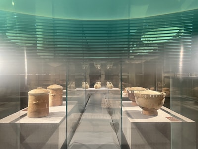 An exhibit of ancient pottery is displayed inside a transparent glass case. The pottery includes two large cylindrical jars and several round bowls, all with an aged, earthy appearance.