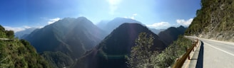 A scenic photo of a winding mountain road surrounded by lush greenery and distant peaks under a clear blue sky.