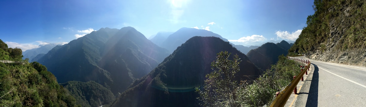 A scenic photo of a winding mountain road surrounded by lush greenery and distant peaks under a clear blue sky.