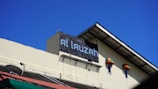 A building with a large, prominent sign displaying the text 'Al lauzah' is shown in the center. The building exterior is cream-colored with a black-tiled sign area. Two small windows with colorful awnings are visible below the sign. A satellite dish is mounted on the left side of the roofline. The sky above is clear and bright blue.