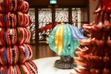 A vibrant and colorful art installation featuring large, textured sculptures with intricate patterns. The central sculpture in turquoise and blue displays circular motifs with text written on it. Surrounding sculptures exhibit bold, geometric designs in reds, pinks, greens, and blues. The background includes a detailed wall art with abstract black and white patterns.
