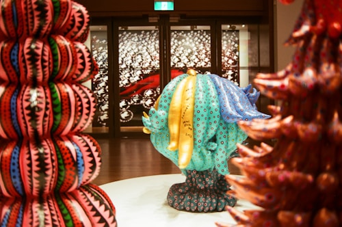 A vibrant and colorful art installation featuring large, textured sculptures with intricate patterns. The central sculpture in turquoise and blue displays circular motifs with text written on it. Surrounding sculptures exhibit bold, geometric designs in reds, pinks, greens, and blues. The background includes a detailed wall art with abstract black and white patterns.