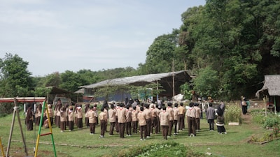 A large group of students in uniform stands in a circular formation on a grassy clearing, surrounded by lush greenery and trees. In the background, there are rustic structures, including a thatched hut and a simple building with a corrugated metal roof. A playground swing set is visible on one side.