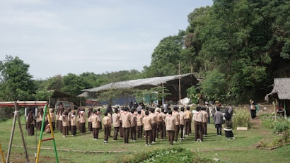 A large group of students in uniform stands in a circular formation on a grassy clearing, surrounded by lush greenery and trees. In the background, there are rustic structures, including a thatched hut and a simple building with a corrugated metal roof. A playground swing set is visible on one side.