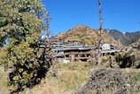 A rural village is nestled among hilly terrain, with rustic houses and wooden structures surrounded by trees and dry vegetation. The sky is clear and bright, indicating a sunny day.