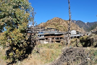 A rural village is nestled among hilly terrain, with rustic houses and wooden structures surrounded by trees and dry vegetation. The sky is clear and bright, indicating a sunny day.