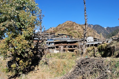 A rural village is nestled among hilly terrain, with rustic houses and wooden structures surrounded by trees and dry vegetation. The sky is clear and bright, indicating a sunny day.