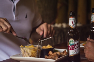 Two people are dining together, with one person holding a knife and fork over a plate. The plate contains fries in a small wire basket and a serving of cooked meat. On the table are two bottles of beer with 'Victoria' labels.