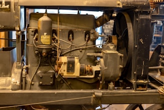 An antique engine with visible components such as gears, pipes, and a cylindrical tank. The engine has a worn, industrial appearance and is mounted on a metal frame.