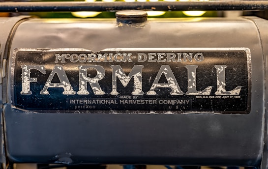 A close-up view of an old Farmall tractor logo. The sign is weathered with visible metal wear and rust, giving a vintage industrial feel. It prominently displays the words 'McCormick Deering Farmall', with smaller text indicating it was made by the International Harvester Company in Chicago, USA.