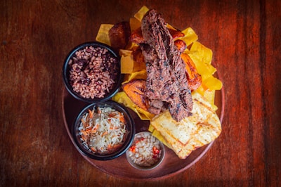 A wooden table holds a rustic plate filled with a variety of foods. In the center, there are grilled beef strips accompanied by vibrant plantain slices, yellow fried slices, and grilled cheese. Surrounding this are small bowls filled with rice and beans, a cabbage and carrot coleslaw, and a separate spicy vegetable mixture. The presentation is colorful and appetizing.