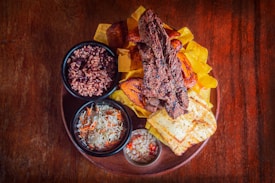 A wooden table holds a rustic plate filled with a variety of foods. In the center, there are grilled beef strips accompanied by vibrant plantain slices, yellow fried slices, and grilled cheese. Surrounding this are small bowls filled with rice and beans, a cabbage and carrot coleslaw, and a separate spicy vegetable mixture. The presentation is colorful and appetizing.