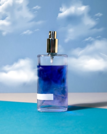 A glass spray bottle filled with a blue liquid stands against a backdrop of a sky with fluffy white clouds. The bottle has a metallic spray nozzle and is placed on a turquoise surface.