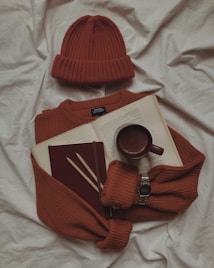 A cozy arrangement featuring a rust-colored knit sweater and matching beanie on a soft white fabric. Placed on the sweater is an open book, a maroon notebook, two wooden pencils, and a brown mug. A silver digital watch is also visible, adding a subtle metallic touch.