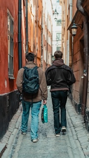 A narrow cobblestone alleyway flanked by tall, warmly colored buildings. Two people are walking away from the camera, one carrying a backpack and the other holding a turquoise bag. The alleyway is lit by a few hanging lamps.