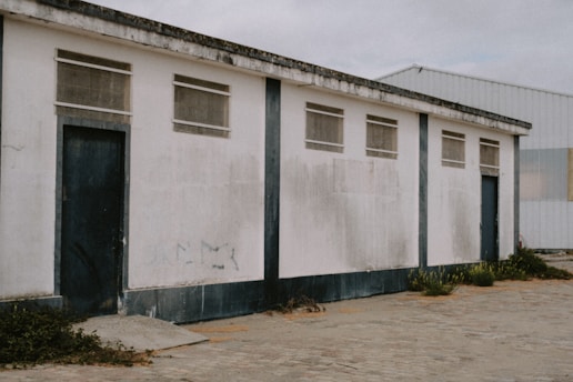 A long, rectangular white building with a flat roof. The exterior walls are weathered, showing signs of wear and dirt. The building has four small, high-set windows and two large, dark blue doors. There is some overgrown vegetation along the base of the building and cracks in the paved ground in front of it. Another building made of corrugated metal is visible in the background.