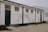A long, rectangular white building with a flat roof. The exterior walls are weathered, showing signs of wear and dirt. The building has four small, high-set windows and two large, dark blue doors. There is some overgrown vegetation along the base of the building and cracks in the paved ground in front of it. Another building made of corrugated metal is visible in the background.
