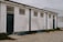 A long, rectangular white building with a flat roof. The exterior walls are weathered, showing signs of wear and dirt. The building has four small, high-set windows and two large, dark blue doors. There is some overgrown vegetation along the base of the building and cracks in the paved ground in front of it. Another building made of corrugated metal is visible in the background.