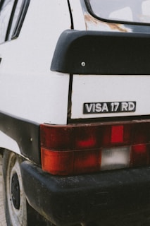 Close-up view of the rear section of a white car, focusing on the taillight and a badge labeled 'VISA 17 RD'. The car appears to have some rust and wear. The image highlights the simple and classic design elements.