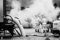 Two workers wearing safety vests stand near a large vehicle and equipment emitting smoke or steam. Traffic cones are placed around them on a cobblestone street. The scene suggests construction or utility work.