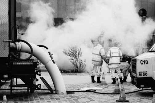 Two workers wearing safety vests stand near a large vehicle and equipment emitting smoke or steam. Traffic cones are placed around them on a cobblestone street. The scene suggests construction or utility work.