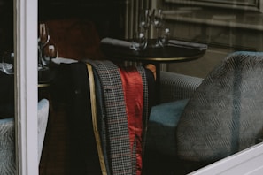Inside a cozy and elegant restaurant, several wine glasses are neatly arranged on round tables, accompanied by folded napkins. A patterned jacket, featuring houndstooth and red satin, is draped over a soft, upholstered armchair with a blue and white design.