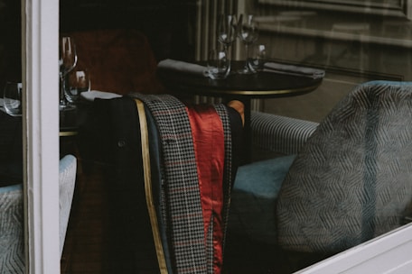 Inside a cozy and elegant restaurant, several wine glasses are neatly arranged on round tables, accompanied by folded napkins. A patterned jacket, featuring houndstooth and red satin, is draped over a soft, upholstered armchair with a blue and white design.